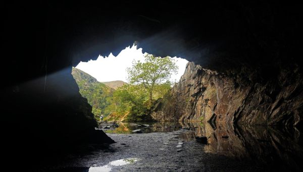 A view from deep within a cave, looking out into a bright, hilly and green landscape in a valley